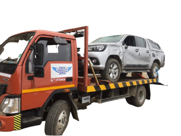 A tow truck from a towing company carrying a damaged silver pickup truck on its flatbed, featuring a logo and contact details on the side.